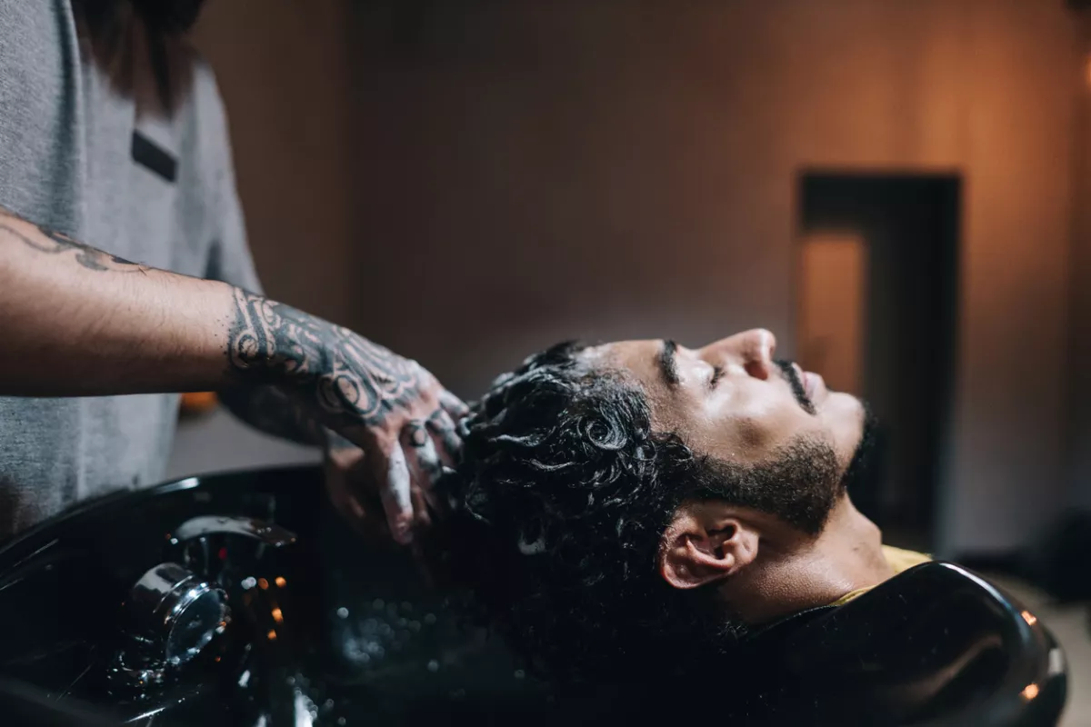 Foto de un hombre de barba al que le lavan el cabello rizado, negro y abundante en una barbería.