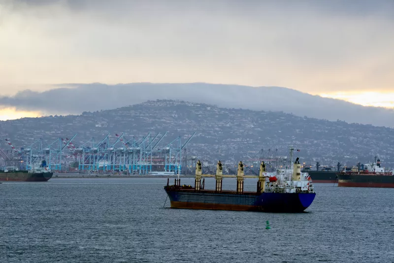The City Of Ensenada, Mexico In The Background With A Barge Passing By In The Commercial Shipping Harbor