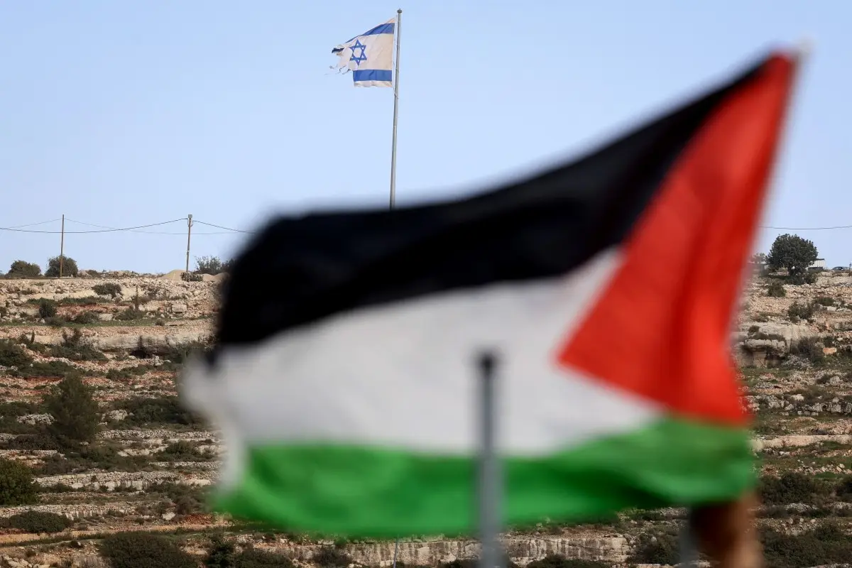 Esta foto tomada desde la aldea de Umm Safa, al norte de Ramallah en la Cisjordania ocupada, muestra una bandera palestina (en primer plano) y una bandera israelí ondeando en la cima de una colina con vistas al pueblo el 16 de febrero de 2026. El gobierno de Israel ha aprobado un proceso para registrar la tierra en Cisjordania como "propiedad estatal", lo que provocó la condena de las naciones árabes y los críticos que dijeron que aceleraría la anexión del territorio palestino.