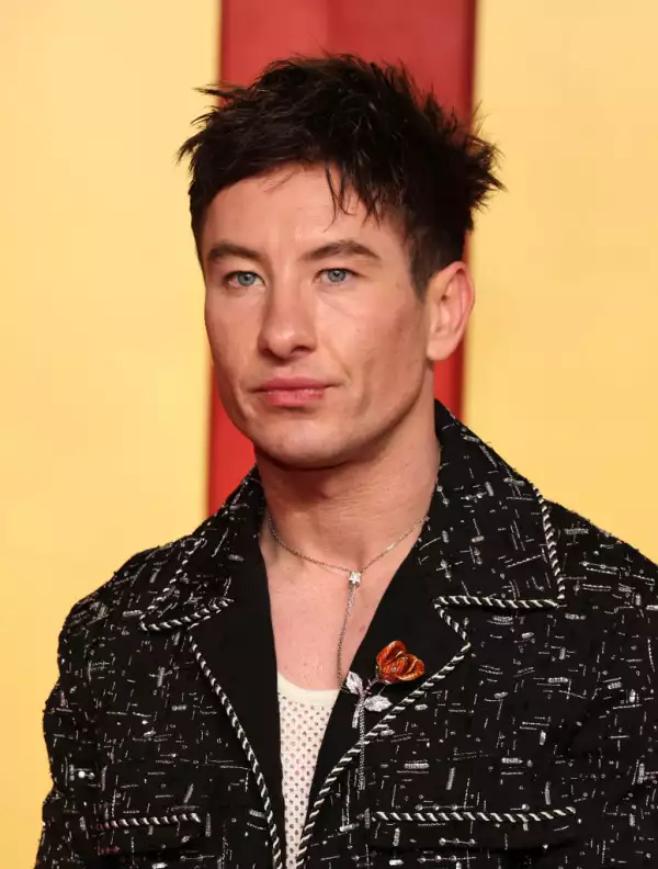 Foto de Barry Keoghan utilizando un traje negro con un collar delicado y delgado largo y un broche de flor roja