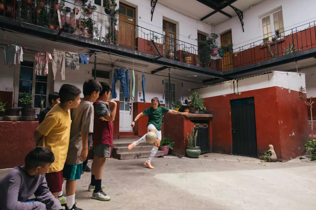 La futbolista Jacquie Ovalle pateando un balón mientras es observada por algunos niños en una vecindad. NIKE Piérdelo todo, gánalo todo.