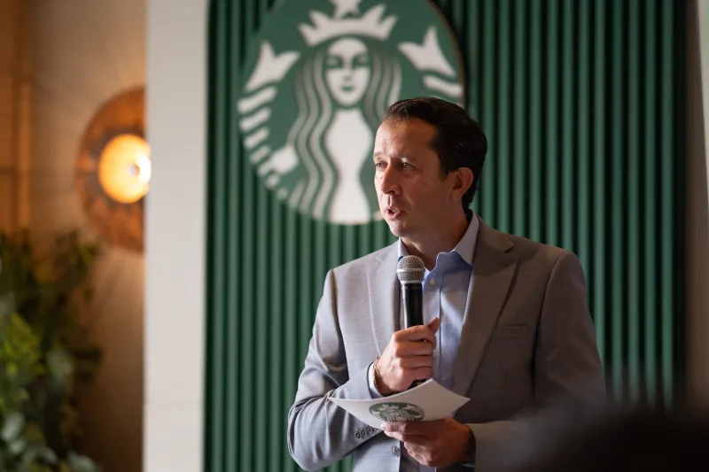 Jaime Vásquez, director de Starbucks, en traje gris con camisa azul. Sostiene un micrófono en la mano izquierda y una tarjeta en la mano derecha. Al  fondo está una pared color verde en la que sobre sale el logitipo de Starbucks, que es una sirena con dos colas en color verde.