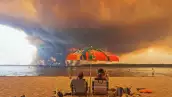Los bañistas se sientan en la playa de Moutchic en Lacanau, al suroeste de Francia, el 24 de julio de 2026, mientras las nubes de humo se elevan al cielo a partir de un incendio forestal.