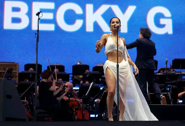 Foto de Becky G con un vestido de dos piezas cantando en Coachella 2025, a su espalda se ve a Gustavo Dudamel dirigiendo a la orquesta filarmónica de Los Ángeles.