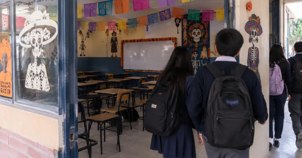 Cuatro jóvenes estudiantes con mochila al hombro saliendo de un salón de clases adornado con papel picado y catrinas.jpg