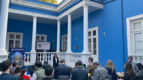 Ventanales y columnas de una casa azul con blanco. Personas sentadas en el exterior frente a una mujer con micrófono.jpg