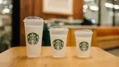 Tres vasos blancos de Starbucks de distintos tamaños, con tapas transparentes y el logotipo verde, colocados sobre una mesa de madera dentro de una cafetería.