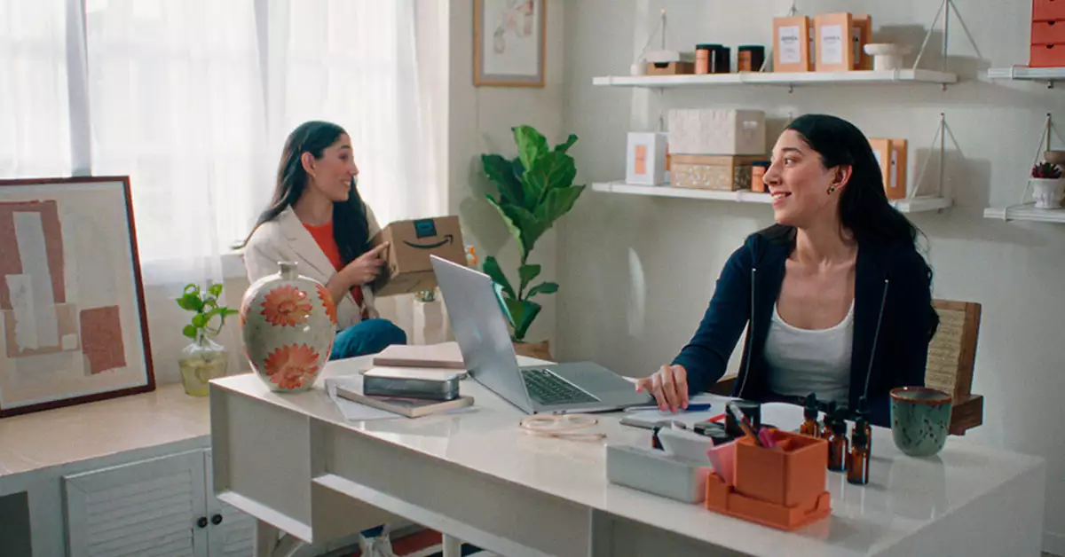 Dos mujeres, sentadas, detrás de un escritorio blanco, en una oficina blanca, una de las chicas de lado izquierdo, sostiene una caja con logotipo de amazon
