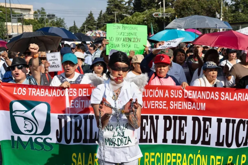 Trabajadores del IMSS protestando en una calle, varios sostienen una manta larga en la que exigen solución a sus demandas salariales; enfrente una persona con el uniforme blanco, las manos amarradas y la boca y los ojos tapados. 