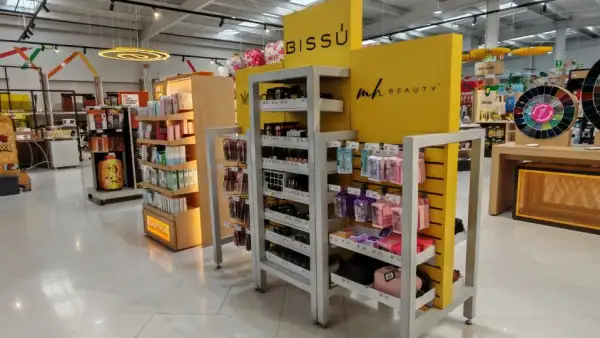nterior de una tienda OfficeMax con un exhibidor amarillo de Bissú y productos de maquillaje y belleza distribuidos en estantes. Al fondo se observan otras áreas de venta y pasillos de la sucursal. La imagen muestra la presencia de categorías de cuidado personal dentro del establecimiento.