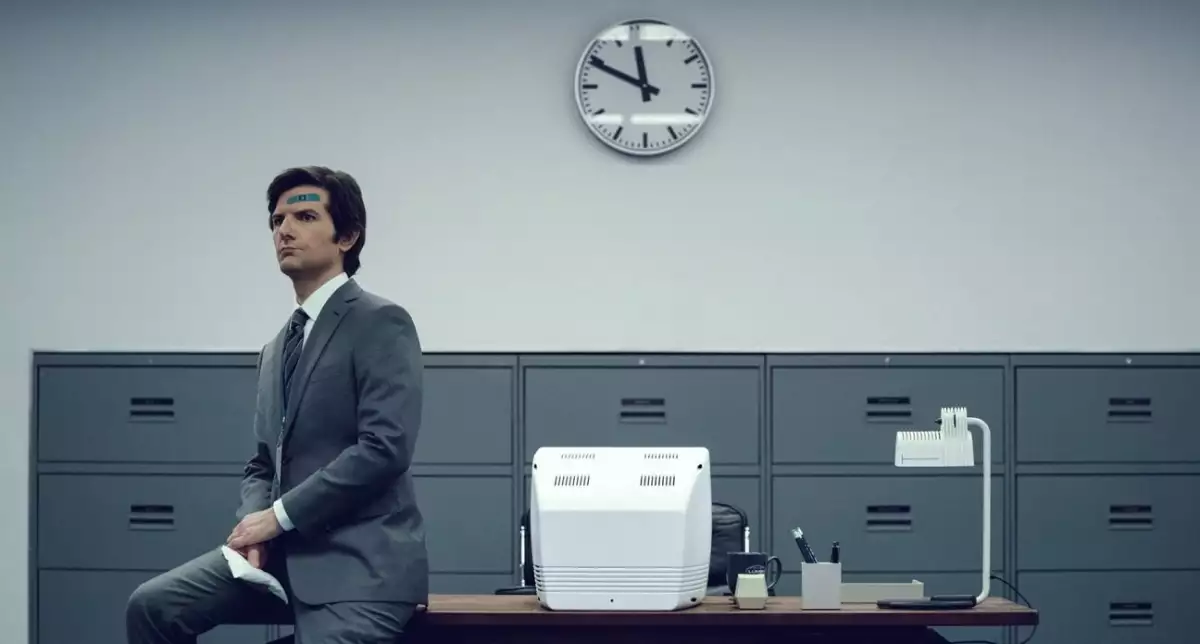 Foto de Adam Scott como Mark, personaje de 'Severance' sentado en la esquina de un escritorio vestido de gris en una oficina llena de archiveros grises.