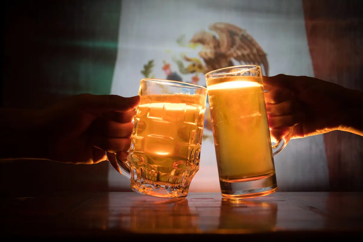 (Dos personas chocando tarros de cerveza frente a una bandera de México)