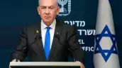 Benjamin Netanyahu, un hombre blanco de 74 años. Cabello blanco canoso, viste un traje negro, una camisa blanca y corbata azul vibrante. Porta una expresión seria. Detrás de él, una bandera de Israel.