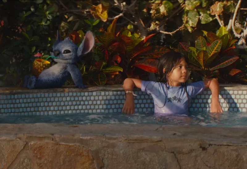 Foto de una escena de la película live action Lilo y Stitch, con Lilo en una alberca y Stitch sentado a su lado tomando una una bebida en una piña.