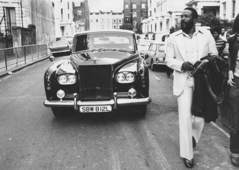 Foto en blanco y negro de Marvin Gaye con un traje blanco caminando por la calle, detrás de él su auto Rolls Royce.