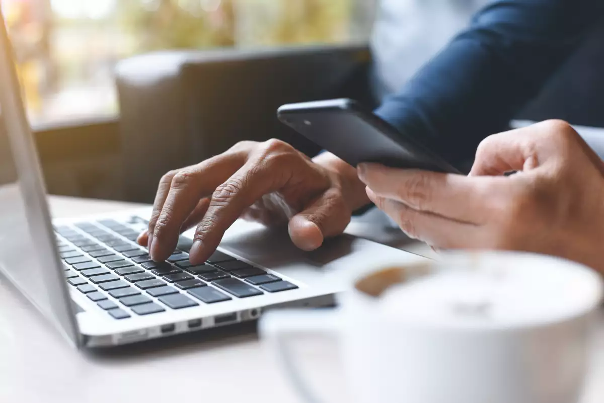 Cierre de un hombre casual o trabajador independiente trabajando en una laptop, sosteniendo un teléfono móvil inteligente con café en una cafetería 