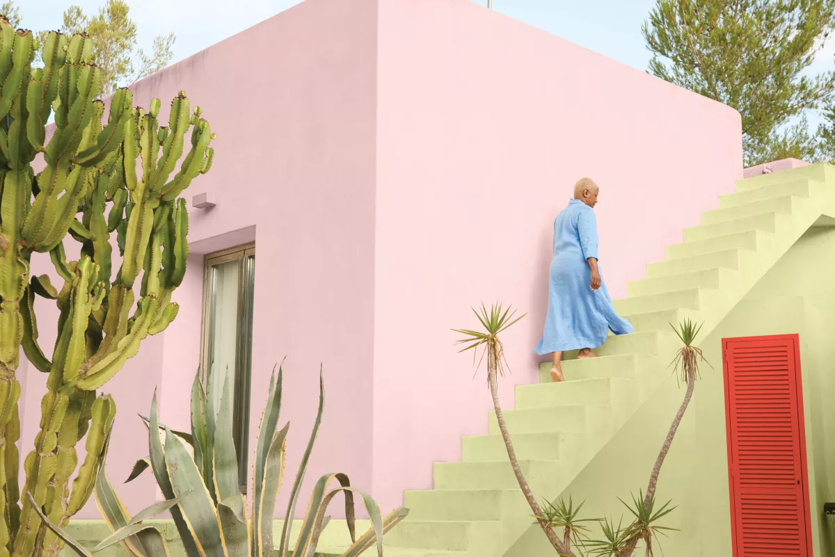 Una mujer asciende por unas escaleras en un edificio pintado con color plasma, un tono rosado. Al frente se observan unas plantas.