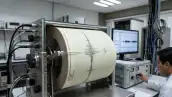 Un sismógrafo leyendo señales de algún movimiento telúrico, de fondo hay una persona monitoreando los datos en una pantalla. 