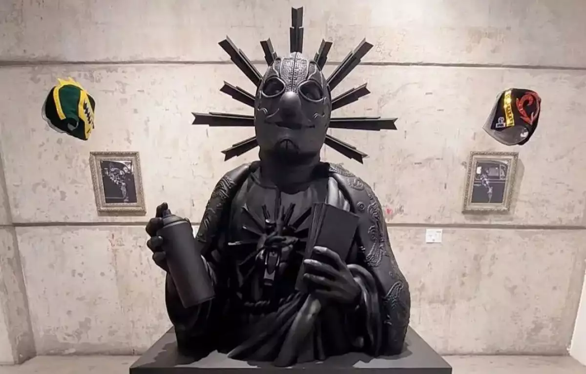 Foto de una escultura negra de un busto de luchador con máscara vestido como de santo que lleva en una mano una botella de pintura en aerosol y en la otra una libreta.