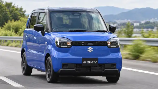 Suzuki e-Sky azul circulando por una carretera, visto de frente, con montañas y vegetación al fondo.