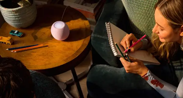 Vista del HomePod mini 2, de Apple, sobre una mesa de centro junto a una mujer dibujando.