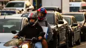 Dos personas viajan en una moto, con otros vehículos de fondo.