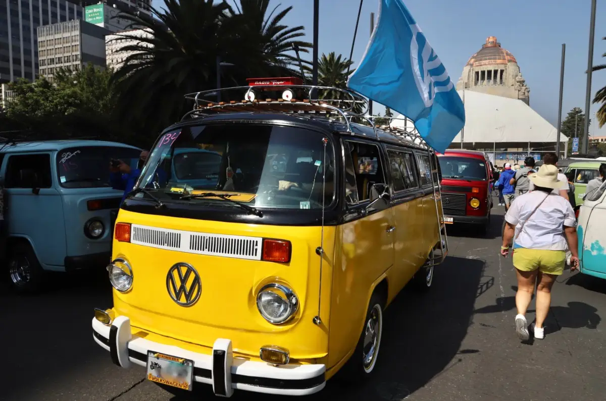 Combi amarilla con bandera de VW transita frente al Monumento a la Revolución.