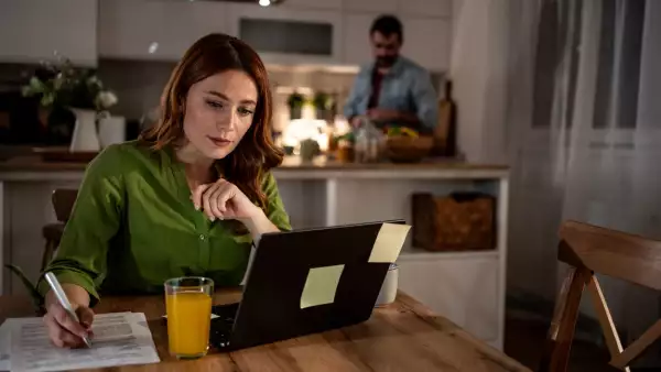 Focused woman working from home while man prepares dinner