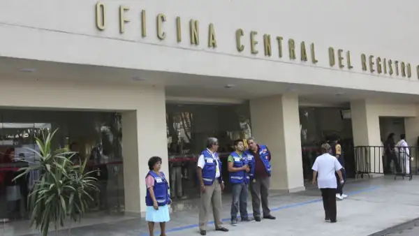 Fachada de edificio color beige con letras de "Oficina Central del registro Civil" y cuatro hombres y una mujer con chaleco azul .jpg