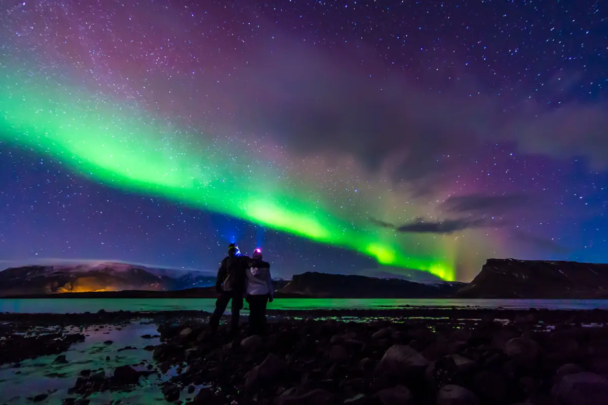 Foto de una pareja observando auroras boreales en Islandia, los tonos de las luces van desde verde intenso hasta rosáceo y morado.