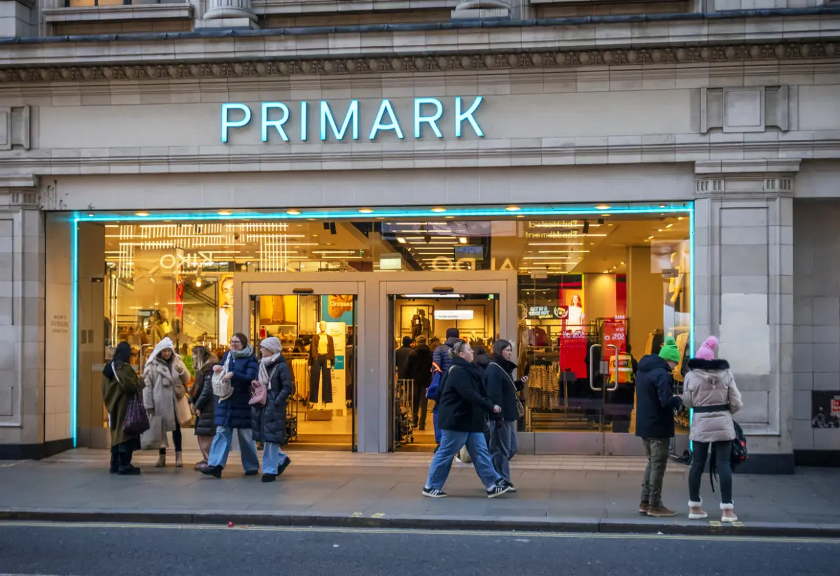 Fachada de una tienda Primark con un letrero de letras azul turquesa iluminadas sobre una entrada amplia. A través de los ventanales se observa el interior de la tienda, con luces cálidas amarillas, exhibidores de ropa y maniquíes. Varias personas con abrigos oscuros y de colores claros caminan o se detienen frente al establecimiento, ubicado en un edificio de piedra gris.