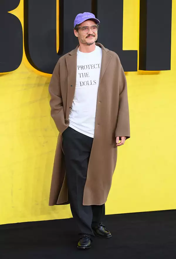 Foto de cuerpo completo de Pedro Pascal con una chaqueta marrón, pantalones oscuros holgados y la playera 'Protect the dolls'