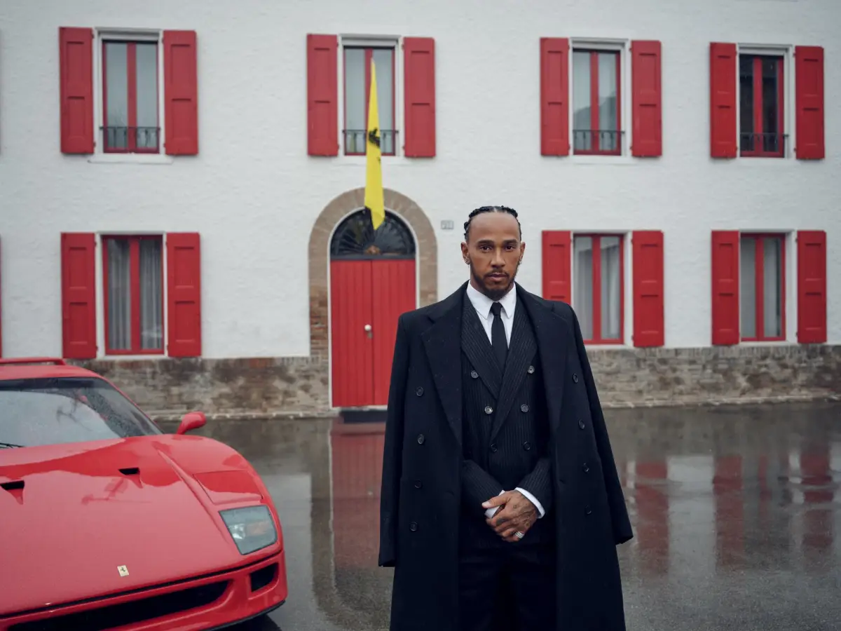 Foto de Lewis Hamilton vestido de traje y abrigo negros, con las manos cruzadas al frente y parado frente a un auto Ferrari color rojo.