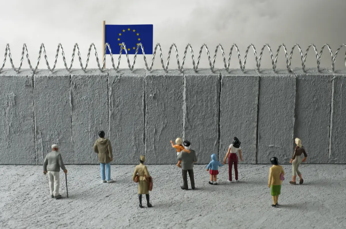 Concepto de crisis de refugiados de la Unión Europea. Personas delante de un muro fronterizo con una bandera de la UE. (Imagen principal del artículo web 'Al igual que Estados Unidos, Europa también endurece sus políticas migratorias'). 