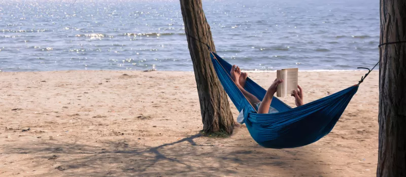 Foto de un hombre en una hamaca leyendo un libro en la playa frente al mar