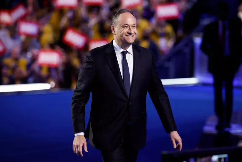 Doug Emhoff, un hombre de 59 años vistiendo un traje negro, camisa blanca y corbata azul. Va caminando sobre un escenario con el fondo de cientas de personas y carteles rojos con su nombre.