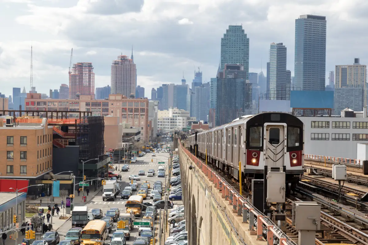 Tren acercándose a la estación de metro elevada en Queens, Nueva York. Los edificios financieros y el horizonte de Nueva York se ven en el fondo, a la izquierda abajo se puede ver una calle concurrida llena de coches en hora punta, cielo nublado, dramático, EE. UU.