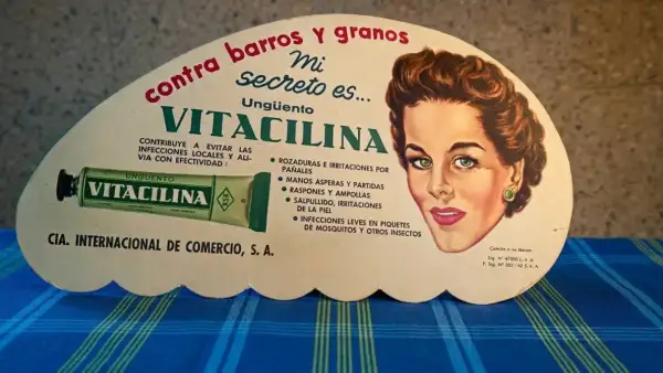 Anuncio publicitario en forma de abanico de Vitacilina. Es de aspecto medio ovalado y en él se ve el rostro de una mujer en los 60, un tubo de Vitacilina y frases como "Mi secreto es..." y el nombre de la marca. 