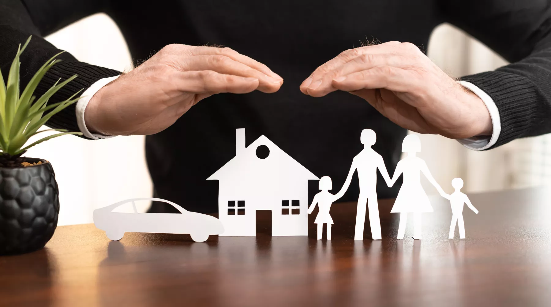 Una imagen que simula protección, ya que hay una figura de papel de un auto, una casa y una familia que son cubiertas por las manos de una persona.