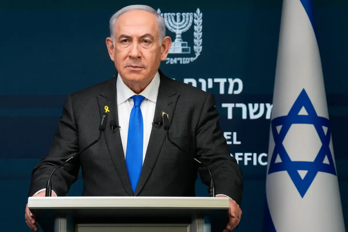 Benjamin Netanyahu, un hombre blanco de 74 años. Cabello blanco canoso, viste un traje negro, una camisa blanca y corbata azul vibrante. Porta una expresión seria. Detrás de él, una bandera de Israel.