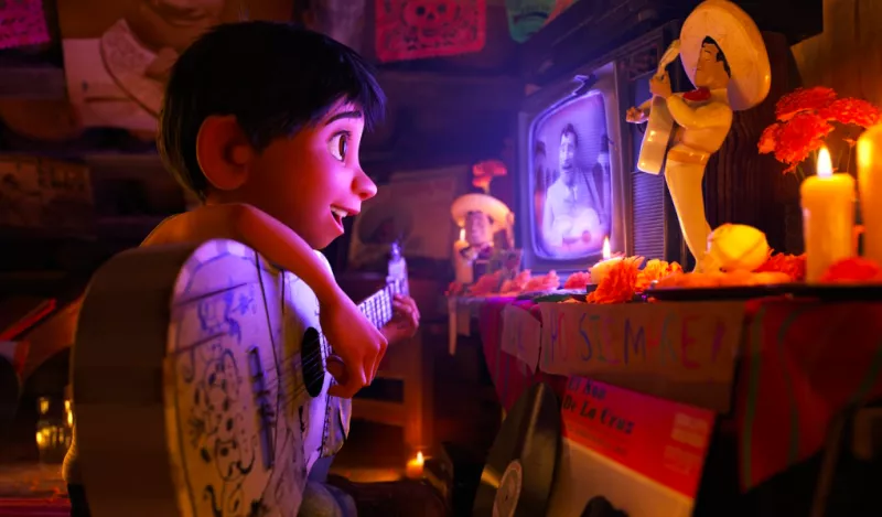 Foto de una escena de la película animada de Pixar, Coco, con el pequeño Miguel tocando su guitarra viendo emocionado la televisión.