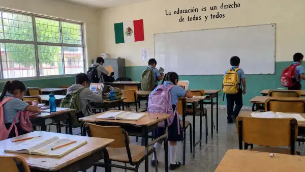 Bancas de color café enfrente de un pizarrón en blanco en un salón de clases con siete alumnos con mochilas en la espalda.jpg