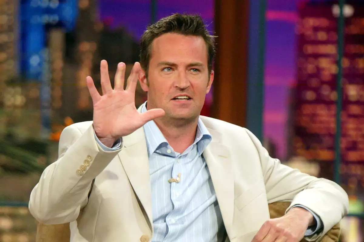 Foto de Matthew Perry en 2004 durante una entrevista en un programa de televisión con un saco color calor y camisa azul cielo, levantando una mano.