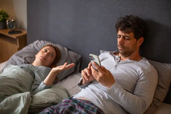 Man ignoring woman in bed while phubbing using smartphone
