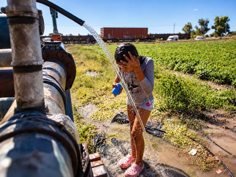 Niña se moja con agua que sale de una tubería en un campo agrícola, mientras un tren de carga circula al fondo.