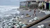 Costales a la orilla de una playa que luce con nubes. Un hombre con bermudas y gorra roja camina frente a un local.jpg