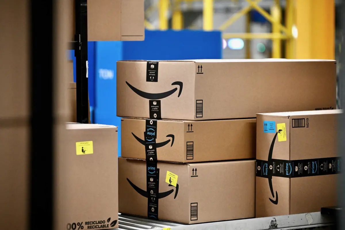 Paquetes de cartón de Amazon, algunos con cinta de embalaje de Prime y etiquetas de envío, apilados sobre una banda transportadora dentro de un centro logístico, con estructuras y maquinaria industrial al fondo.