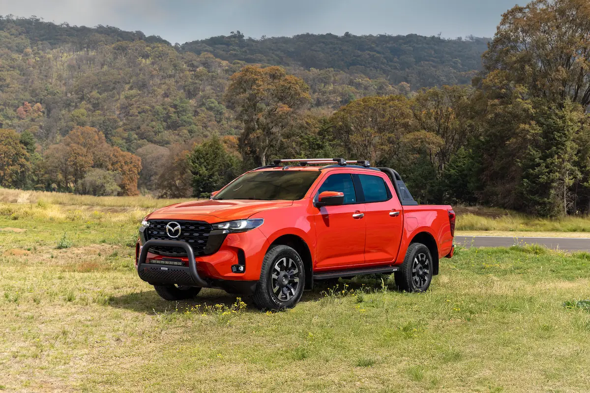 Imagen de la nueva pickup Mazda Bt-50 color rojo, estacionada en un paraje con pasto verde con un bosque y montañas de fondo.