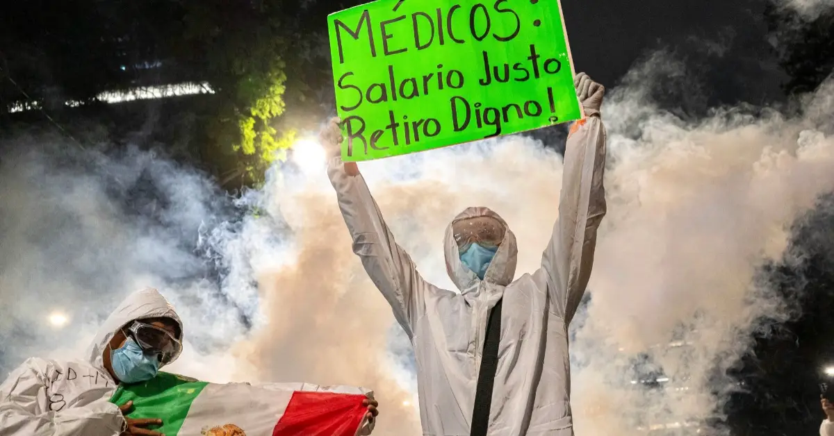 Dos personas con bata blanca y cubrebocas y humo detrás con un cartel que dice: Médicos: salario justo.jpg