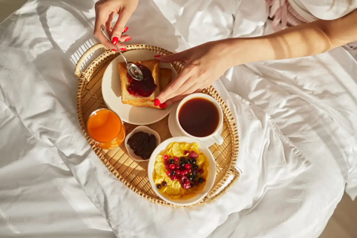 Desde arriba de una mujer sentada en la cama y esparciendo mermelada dulce sobre una tostada mientras desayunaba en el cuarto en la mañana soleada